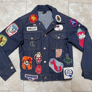 May include: A dark blue denim jacket with a variety of colorful patches sewn on. The patches include cartoon characters, animals, and logos. The text "SHELLEY" is on one of the patches.