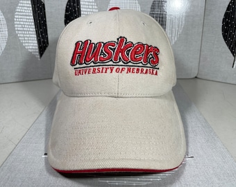 May include: A beige baseball cap with red and black embroidery that reads "Huskers University of Nebraska".