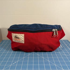 May include: A red and navy blue Kelty fanny pack with a black zipper and silver zipper pulls. The pack has a white rectangular tag with the Kelty logo and text. The fanny pack is on a blue grid surface.