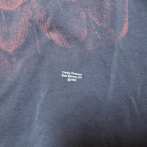 May include: Navy blue t-shirt with a faded red design. The text "Trinity Products San Marcos, CA &copy;1993" is printed in white. The fabric has a textured appearance.