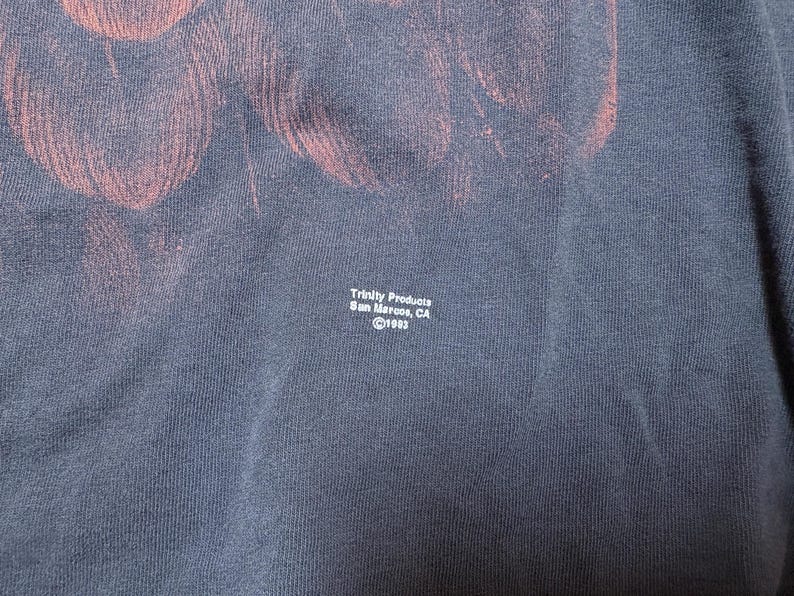 May include: Navy blue t-shirt with a faded red design. The text "Trinity Products San Marcos, CA &copy;1993" is printed in white. The fabric has a textured appearance.