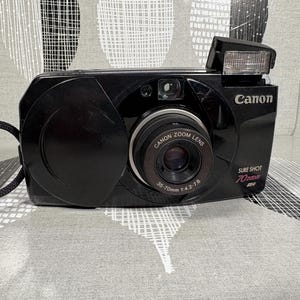 May include: A black Canon Sure Shot 70 Zoom camera with a zoom lens and built-in flash. The camera has a black strap and the Canon logo is visible. The lens has the text "Canon Zoom Lens 35-70mm 1:4.2-7.8".
