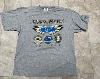 Peut inclure: Un t-shirt gris avec un motif Star Wars Episode I. Le motif représente le chasseur de la Fédération du Commerce, le chasseur de Naboo et le chasseur N-1. Le texte "STAR WARS" est imprimé en lettres vertes et noires au-dessus du motif. Le texte "EPISODE I" est imprimé en lettres noires en dessous du motif.