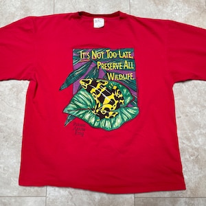 Peut inclure: T-shirt rouge à manches courtes avec une grenouille empoisonnée à flèche verte, jaune et noire sur une feuille. Le t-shirt porte l'inscription "It's Not Too Late. Preserve All Wildlife."