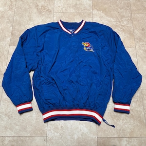 May include: A blue pullover jacket with a V-neck collar and long sleeves. The jacket has red and white striped cuffs and a matching hem. A logo of a Jayhawk is embroidered on the chest. The jacket is made of a lightweight, crinkled fabric.