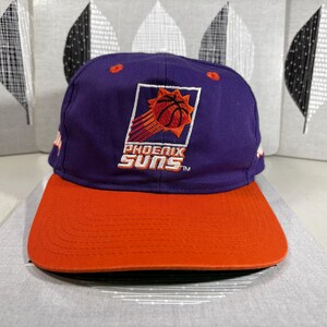 Könnte beinhalten: Eine lila-orangefarbene Phoenix Suns Baseballkappe. Auf der Kappe befindet sich das Logo des Teams mit einem Basketball- und Sonnendesign. Der Schirm ist orange und die Krone lila.