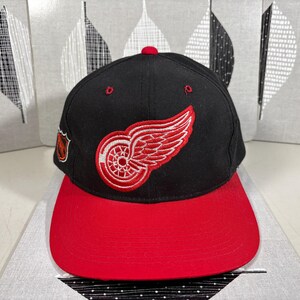 Puede incluir: Gorra de béisbol negra y roja con el logotipo de los Red Wings de Detroit. La gorra tiene una visera roja y una corona negra con el logotipo de la rueda alada del equipo bordado en rojo y blanco. Un pequeño parche con el logotipo del equipo está en el lateral.
