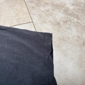 May include: Close-up of a dark grey t-shirt. The fabric has a slightly faded appearance. The edge of the shirt is visible, showing the hem and stitching details. The background is a light-coloured, textured surface.