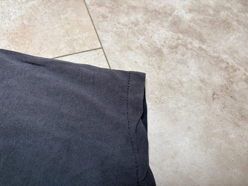 May include: Close-up of a dark grey t-shirt. The fabric has a slightly faded appearance. The edge of the shirt is visible, showing the hem and stitching details. The background is a light-coloured, textured surface.