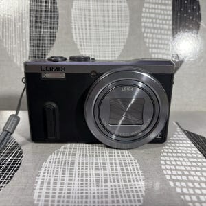 May include: A black and silver Lumix digital camera with a Leica lens. The camera has a retractable lens and a gray wrist strap. The camera is on a patterned surface.