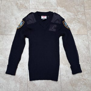Könnte beinhalten: Marineblauer V-Ausschnitt-Pullover mit langen Ärmeln und gerippten Bündchen. Mit Schulteraufnähern und Aufnähern mit einem gold-blauen Emblem. Ein Etikett ist am Halsausschnitt sichtbar. Der Pullover ist aus dickem Strickmaterial.