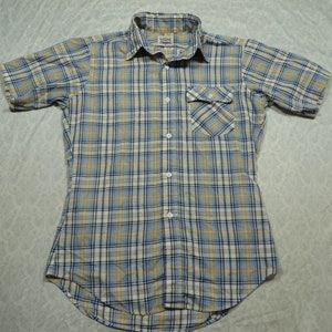 Puede incluir: Una camisa de manga corta a cuadros con botones con un patrón azul claro, beige y negro. La camisa tiene un solo bolsillo en el pecho y un cuello abotonado.