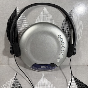 Könnte beinhalten: Ein silberner tragbarer CD-Player mit kreisförmigem Design und angeschlossenem schwarzem kabelgebundenem Headset. Das Gerät verfügt über eine Reihe kleiner runder Steuertasten auf der rechten Seite und ein blaues, durchscheinendes Feld unten mit dem RCA-Logo in Weiß.