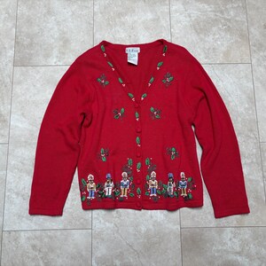 Op de afbeelding: Een rode cardigan met een V-hals en knoopsluiting. De trui is versierd met geborduurde hulst en notenkrakerfiguren. Een feestelijk kledingstuk voor de feestdagen.