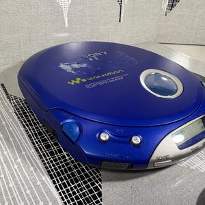 May include: A blue Sony Walkman portable CD player featuring a circular viewing window, a silver digital display panel, and control buttons. The device is labeled with the Sony logo, Walkman branding, and ESP MAX CD-R/RW text on the top surface.