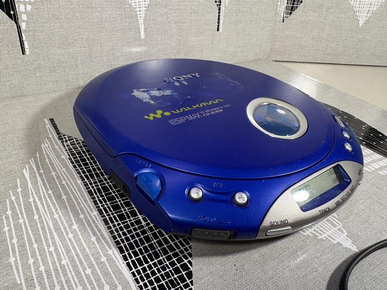 May include: A blue Sony Walkman portable CD player featuring a circular viewing window, a silver digital display panel, and control buttons. The device is labeled with the Sony logo, Walkman branding, and ESP MAX CD-R/RW text on the top surface.