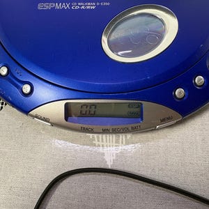 May include: A blue portable CD player with a circular viewing window and a silver control panel. The device features the text ESP MAX CD WALKMAN D-E350 CD-R/RW, along with buttons labeled SOUND, MENU, TRACK, MIN, SEC, VOL, and BATT, and a digital display.