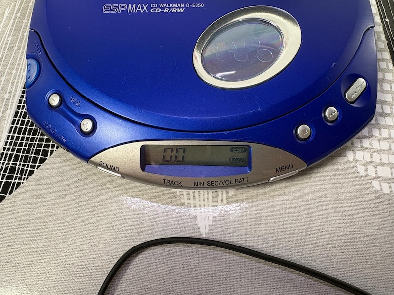 May include: A blue portable CD player with a circular viewing window and a silver control panel. The device features the text ESP MAX CD WALKMAN D-E350 CD-R/RW, along with buttons labeled SOUND, MENU, TRACK, MIN, SEC, VOL, and BATT, and a digital display.