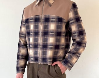 May include: A zip-up jacket with a plaid pattern in navy, beige, and cream. The jacket features a solid tan panel across the shoulders and collar. The model is wearing brown pants. The jacket has a silver zipper.