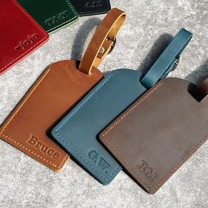 Könnte beinhalten: Eine Sammlung von Leder-Gepäckanhängern in verschiedenen Farben, darunter Rot, Grün, Schwarz, Braun und Blau. Jeder Anhänger hat einen Riemen und personalisierten Text, wie Namen oder Initialen, auf der Vorderseite.