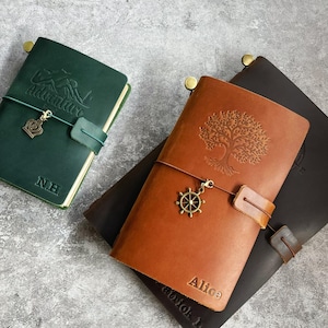 Könnte beinhalten: Drei Lederjournale in Grün, Braun und Dunkelbraun. Das grüne Journal hat das Wort "Adventure" auf dem Einband geprägt, darunter die Initialen "N.H.". Das braune Journal zeigt ein Baumdesign und den Namen "Alice". Jedes Journal hat einen Anhänger und einen Riemen.