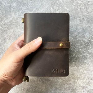 Może przedstawiać: Ciemnobrązowy skórzany notatnik z wytłoczonym na okładce imieniem "Mills". Notatnik jest zabezpieczony skórzanym paskiem i mosiężnym zapięciem na guziki. Notatnik jest trzymany w dłoni.