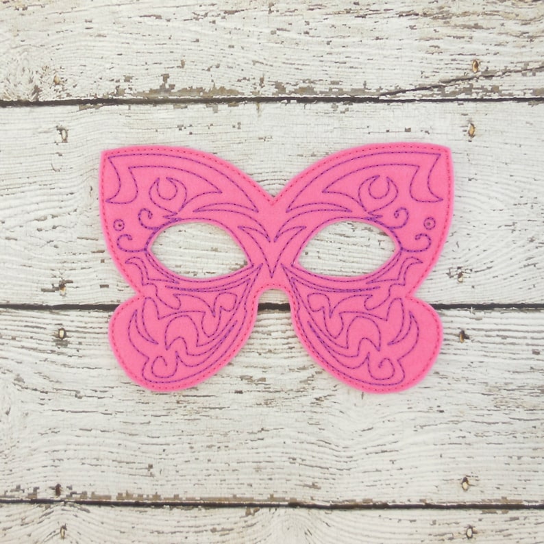 May include: A pink felt masquerade mask with a purple intricate design. The mask has two eye holes and is shaped like a butterfly.