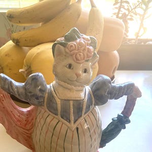 May include: A ceramic teapot shaped like a cat wearing a bonnet and dress. The teapot is primarily white with blue, pink, and green accents. The cat holds a pink shawl and a green umbrella. The teapot has a striped pattern and a floral design on the bonnet.