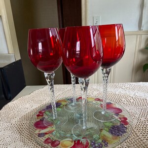 May include: Four red wine glasses with clear twisted stems are displayed on a glass serving tray with a fruit design. The tray is on a crocheted tablecloth.