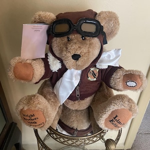 May include: A plush teddy bear wearing a brown aviator hat and goggles, a brown jacket with a white scarf, and brown paws. The bear has the text "Wright Brothers 100th Anniversary" on one paw and "Limited Edition" on the other.