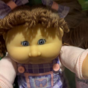 May include: A doll with brown yarn hair styled with purple and white plaid bows. The doll has blue eyes and wears a light pink long-sleeved shirt under purple and white plaid overalls featuring two pink buttons and a central patch with a bow graphic.