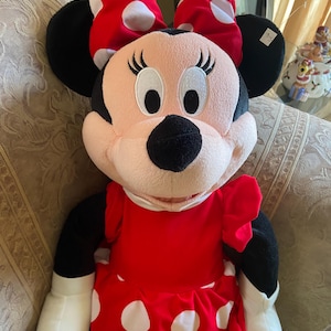 May include: A plush Minnie Mouse doll, featuring a red dress with white polka dots and a matching bow. The doll has black ears, a black nose, and white gloves. The doll is sitting on a patterned surface.