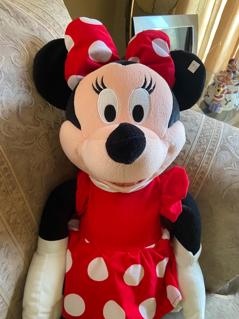 May include: A plush Minnie Mouse doll, featuring a red dress with white polka dots and a matching bow. The doll has black ears, a black nose, and white gloves. The doll is sitting on a patterned surface.