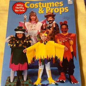 May include: A book titled "Costumes & Props" with a red circle on the cover that says "Nifty, Thrifty, No-Sew". The book features a group of children dressed in costumes, including a fairy, a cowboy, a tiger, a dinosaur, and a chicken.