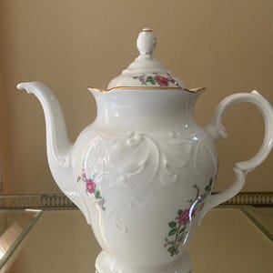 May include: A white porcelain teapot with a lid, spout, and handle. The teapot features embossed floral designs and is decorated with pink roses and green leaves. The rim and lid are trimmed with gold accents. The teapot is sitting on a reflective surface.