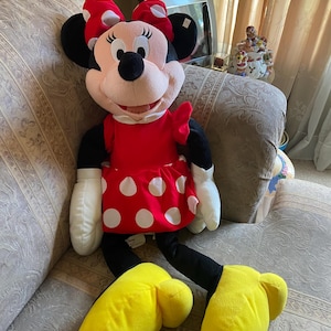 May include: A large Minnie Mouse plush doll. The doll has a red dress with white polka dots, a matching bow, and yellow shoes. The doll's face is pink with black eyes, a black nose, and black ears. The doll is sitting on a patterned couch.