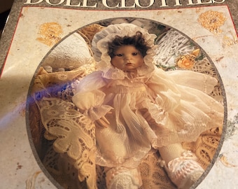 Pode incluir: A capa do livro "The Perfect Book of Doll Clothes" apresenta uma boneca de porcelana vestindo um vestido de renda branca e um chapéu branco. A boneca está sentada em um fundo de tecido de renda. O título do livro está em negrito, em preto. A coleção Vanessa-Ann está escrita em preto na parte inferior da capa.