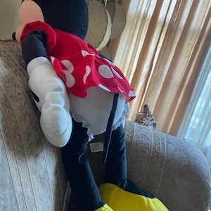 May include: A large plush Minnie Mouse doll. The doll is wearing a red dress with white polka dots, white gloves, and yellow shoes. The doll has black ears and a black head.