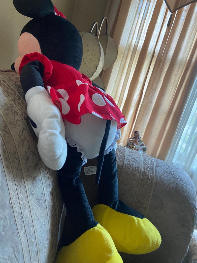May include: A large plush Minnie Mouse doll. The doll is wearing a red dress with white polka dots, white gloves, and yellow shoes. The doll has black ears and a black head.