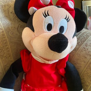 May include: A plush Minnie Mouse doll. The doll has a pink face, black ears, and a red dress with white polka dots. A large red bow with white polka dots sits atop her head. The doll has black arms and a white collar.