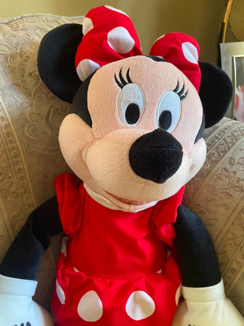 May include: A plush Minnie Mouse doll. The doll has a pink face, black ears, and a red dress with white polka dots. A large red bow with white polka dots sits atop her head. The doll has black arms and a white collar.