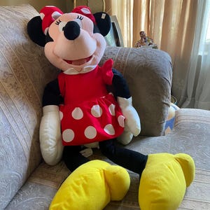 May include: A large Minnie Mouse plush doll. The doll has a red dress with white polka dots, a red bow, and yellow shoes. The doll has black ears, arms, and legs, and white gloves. The doll is sitting on a patterned couch.
