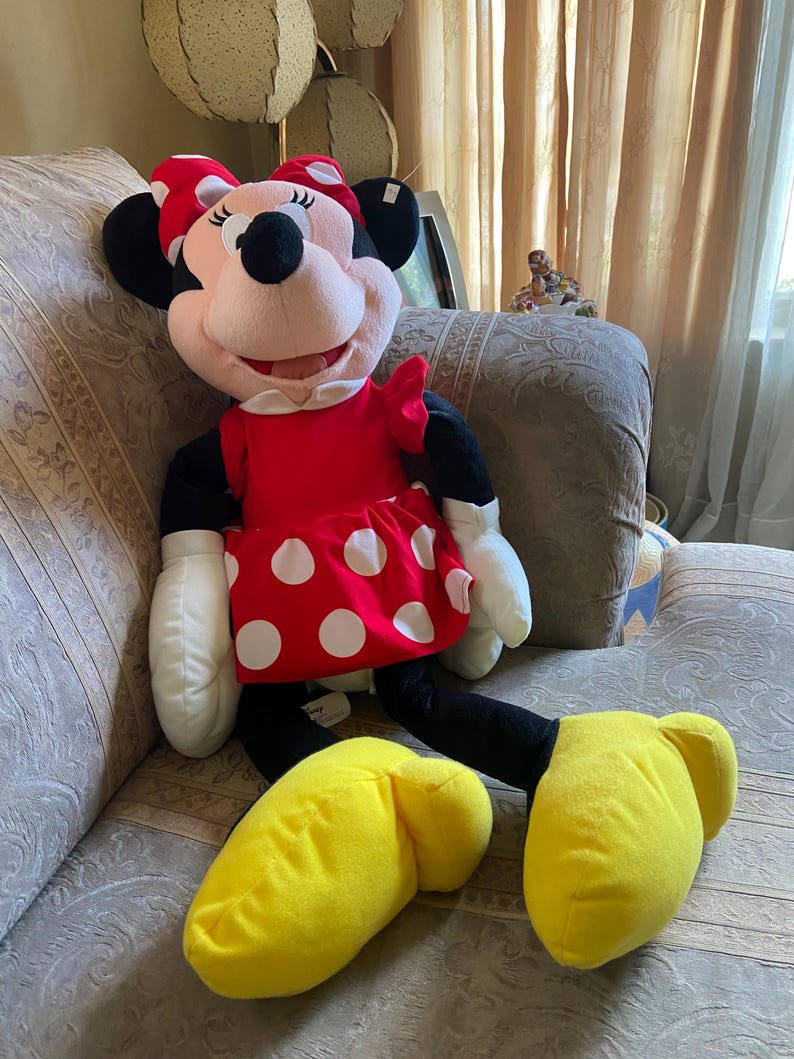 May include: A large Minnie Mouse plush doll. The doll has a red dress with white polka dots, a red bow, and yellow shoes. The doll has black ears, arms, and legs, and white gloves. The doll is sitting on a patterned couch.