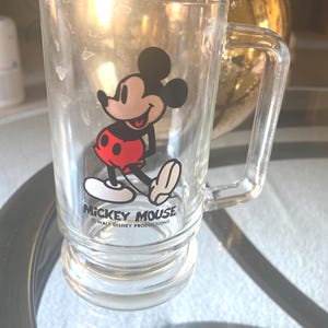 May include: Clear glass mug featuring Mickey Mouse in a classic pose, wearing red shorts and white gloves. The mug has a handle and the words "Mickey Mouse" and "Walt Disney Productions" printed on it. The mug is sitting on a glass surface.