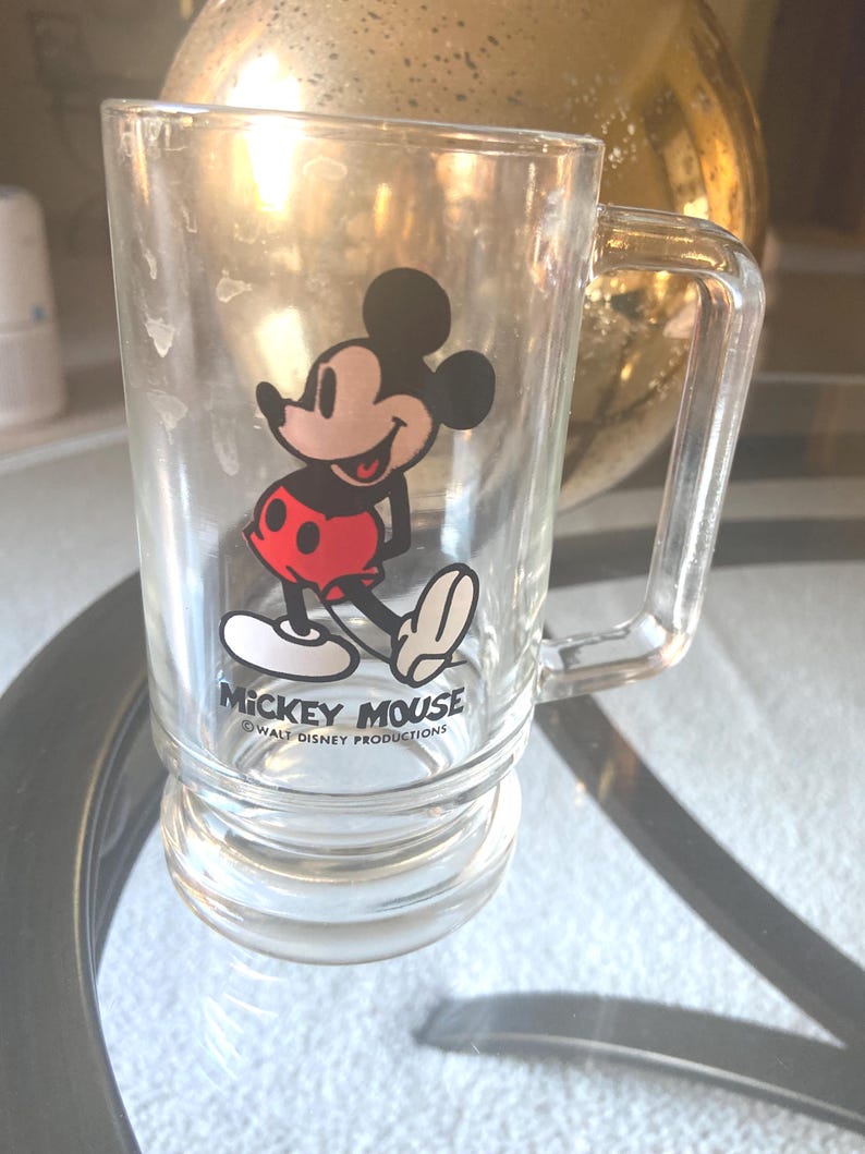 May include: Clear glass mug featuring Mickey Mouse in a classic pose, wearing red shorts and white gloves. The mug has a handle and the words "Mickey Mouse" and "Walt Disney Productions" printed on it. The mug is sitting on a glass surface.