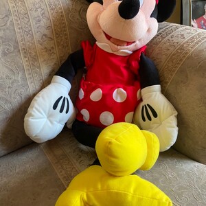 May include: A plush Minnie Mouse doll with a red dress with white polka dots, a matching bow, and white gloves. The doll has black ears, arms, and legs, and yellow feet. The doll is sitting on a patterned sofa.