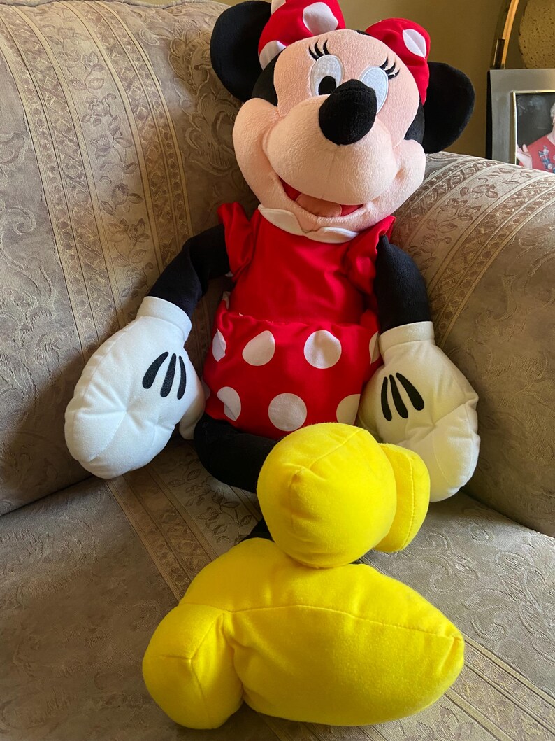 May include: A plush Minnie Mouse doll with a red dress with white polka dots, a matching bow, and white gloves. The doll has black ears, arms, and legs, and yellow feet. The doll is sitting on a patterned sofa.