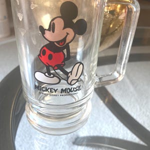 May include: Clear glass mug featuring Mickey Mouse in a classic pose, wearing red shorts and white gloves. The mug has a handle and the words "Mickey Mouse" and "Walt Disney Productions" are printed on the front.