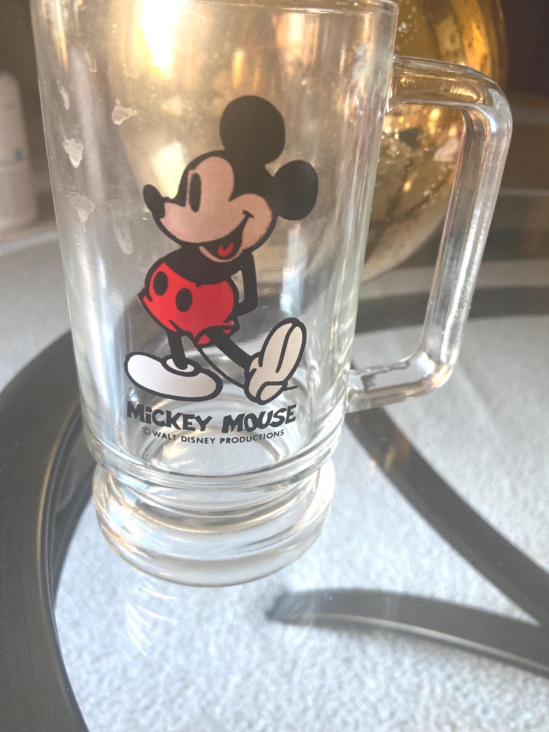 May include: Clear glass mug featuring Mickey Mouse in a classic pose, wearing red shorts and white gloves. The mug has a handle and the words "Mickey Mouse" and "Walt Disney Productions" are printed on the front.