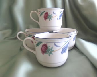 May include: A set of four white ceramic mugs featuring a floral pattern with dark red flowers, green leaves, and blue accents. Each mug has a blue rim, a curved handle, and a stepped base design, arranged in a stacked formation for display.